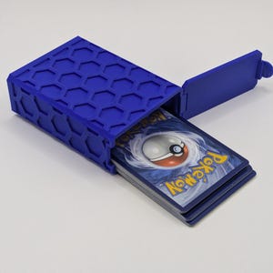 May include: A blue, hexagonal-patterned card storage box with an open lid. The box contains a stack of Pokémon trading cards. The box is made of a solid, durable material and is designed to protect the cards.