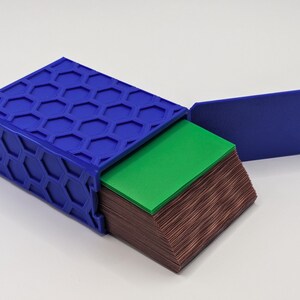 May include: A blue box with a honeycomb pattern on the lid, open to reveal a stack of green and brown cards. The lid is open, revealing the contents. The box is on a white surface.