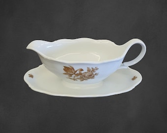 May include: A white porcelain gravy boat with a brown floral design. The boat has a handle and sits on a matching saucer.