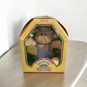 Puede incluir: Un juguete de peluche amarillo Cabbage Patch Kids Koosas en una caja. El juguete lleva un mono azul y un collar marrón con una etiqueta dorada. La caja es amarilla y tiene el logotipo de Coleco y el texto "Una gran mascota para ti y tu Cabbage Patch".