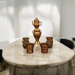 Könnte beinhalten: Ein Set aus sechs Kupfer-Teetassen und einer Kupfer-Teekanne mit aufwendigen floralen Designs auf einem weißen Marmortisch.