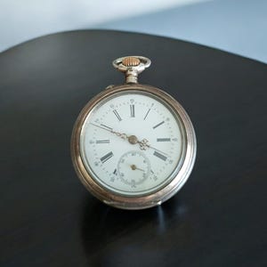 Puede incluir: Un reloj de bolsillo de plata con marcadores de hora en números romanos y una esfera blanca. El reloj tiene una corona de cuerda de color dorado y un pequeño diseño floral decorativo en el centro de la esfera.