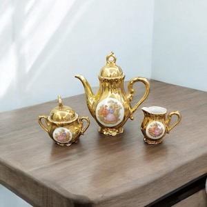 May include: A gold tea set comprising a teapot, sugar bowl, and creamer. Each item has an oval design with a painted scene of figures in historical attire. The set is displayed on a wooden surface.