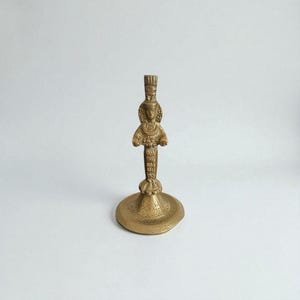 May include: A brass candlestick holder with an ornate design. The candlestick features a detailed figure at the top and a round base with a decorative border. The overall color is a warm, antique gold.
