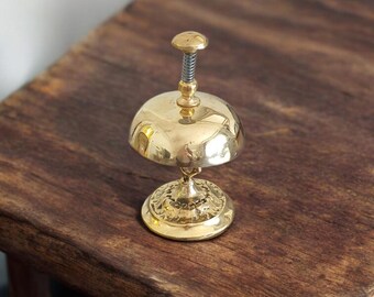 May include: A gold-toned metal service bell with a round top and a decorative base. The bell is on a wooden surface.