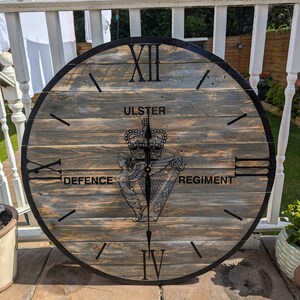 May include: A wooden clock with black Roman numeral markings and a black border. The clock face features a crest with the words "Ulster Defence Regiment" and "Ulster" above the crest.