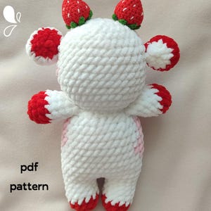 May include: White crocheted stuffed animal with red and pink accents. The animal has a round head with two strawberry-shaped ears and two strawberry-shaped horns. The animal is standing on a white surface. The text "pdf pattern" is visible in the lower left corner of the image.