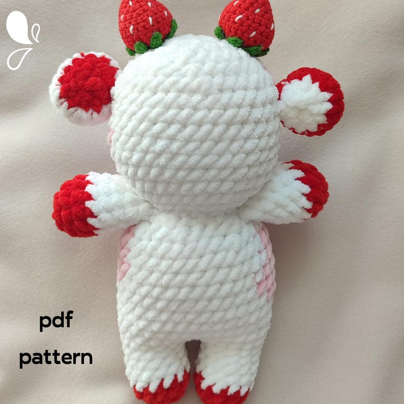 May include: White crocheted stuffed animal with red and pink accents. The animal has a round head with two strawberry-shaped ears and two strawberry-shaped horns. The animal is standing on a white surface. The text "pdf pattern" is visible in the lower left corner of the image.
