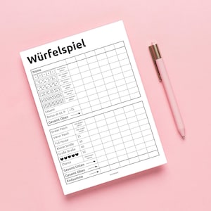 May include: A white dice game score sheet, titled "Würfelspiel" in German, with a grid for recording scores. A pink pen lies on a pink surface beside the sheet. The sheet includes sections for "Name", "Gesamt", and "Chance".