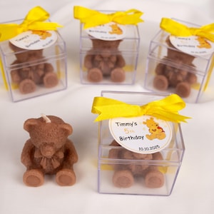 May include: Brown teddy bear candles, some in clear square boxes, are tied with yellow ribbons. Each box has a circular label that reads "Timmy's 5th Birthday 10.10.2025" with a cartoon bear. One candle is outside the box.