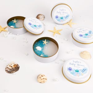 Könnte beinhalten: Weiße Babyparty-Gastgeschenke in Dosen mit goldenen Rändern, mit Meeresmotiven. Jede Dose enthält blaues Gel, eine Muschel und einen Seestern. Die Deckel sind mit dem Text "Sophie's Baby Shower" verziert.