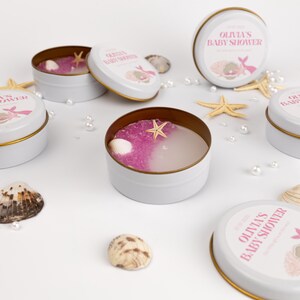 Könnte beinhalten: Weiße Metalldosen mit goldenen Rändern, gefüllt mit rosa und klarem Gel, Muscheln und Seesternen. Die Deckel sind weiß mit dem Text "Olivia's Baby Shower" in Rosa. Dekorative Perlen und Muscheln sind verstreut.