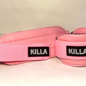 Puede incluir: Correas de tobillo de levantamiento de pesas de color rosa con cierres de velcro ajustables y la palabra "KILLLA" impresa en negro en cada correa.