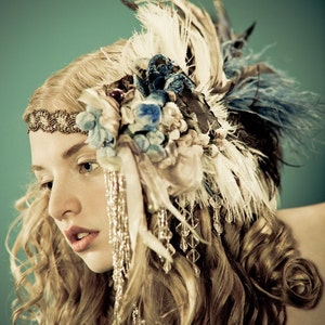 May include: A woman with long, curly blonde hair wears a large, elaborate headpiece made of feathers, beads, and fabric flowers. The headpiece is in shades of white, blue, and brown.