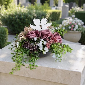Puede incluir: Un arreglo floral con rosas de color rosa polvoriento, pequeñas flores blancas y follaje verde, coronado por dos figuras de ángeles en silueta blanca con halos. El arreglo está contenido en una base circular oscura de perfil bajo.