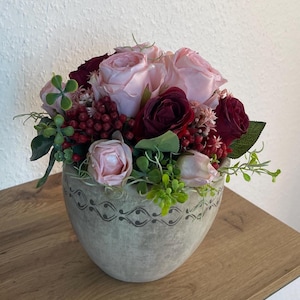 Op de afbeelding: Een bloemstuk met roze en bordeauxrode rozen, rode bessen en groen blad in een decoratieve, lichtgrijze keramische pot. De pot heeft een patroon langs de rand. Het arrangement staat op een houten oppervlak.