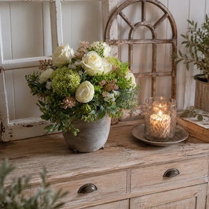 Könnte beinhalten: Ein Blumenarrangement mit cremefarbenen Rosen, grünen Hortensien und verschiedenem Blattwerk in einem runden, verwitterten grauen Topf. Ein gläserner Kerzenhalter mit einer brennenden Kerze steht auf einem kleinen Teller neben dem Blumenarrangement.