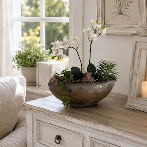 Könnte beinhalten: Eine dekorative Schale mit strukturierter, verwitterter brauner und grauer Oberfläche, die ein künstliches Blumenarrangement enthält. Das Arrangement besteht aus weißen Orchideen, grünen Sukkulenten und kleinem, rosafarbenem Blattwerk.