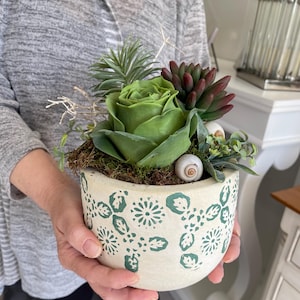 Può includere: Un vaso di ceramica bianco con un motivo floreale verde, riempito con un'arrangiamento di piante grasse che comprende una grande rosa verde, piante grasse più piccole e conchiglie decorative.