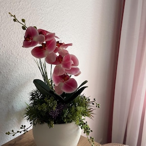 May include: A decorative arrangement featuring pink orchid flowers in a white ceramic pot. The pot is filled with green foliage and small purple flowers. The arrangement sits on a wooden surface, with a pink curtain in the background.