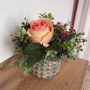 May include: A floral arrangement featuring a large peach-colored rose as the focal point, surrounded by green foliage and small purple berries. The arrangement is in a small, textured, gold-dotted vase, placed on a wooden surface.