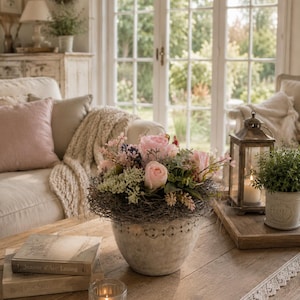 Pode incluir: Um arranjo floral com rosas cor-de-rosa claro, pequenas flores brancas e folhagem verde aninhado num ninho de ramos castanho-escuro, tudo contido numa taça redonda de cerâmica esbranquiçada com um padrão festonado ao longo do rebordo.