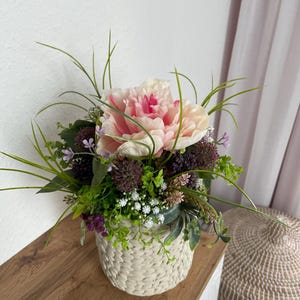 Può includere: Composizione floreale con una grande peonia rosa chiaro al centro, circondata da varie foglie verdi e piccoli fiori viola e bianchi. La composizione è in un cestino intrecciato bianco sporco, posto su una superficie di legno.