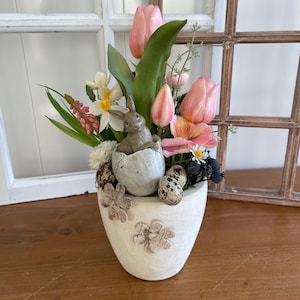 May include: A white ceramic Easter-themed pot with a bunny figurine, speckled eggs, and pink tulips. The pot has a floral design and is filled with greenery and other spring flowers. The arrangement is set on a wooden surface.