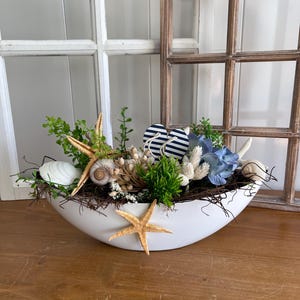 Könnte beinhalten: Eine weiße, oval geformte Dekoschale gefüllt mit einem Küstenarrangement. Es zeigt Muscheln, Seesterne, Grünzeug und blau-weiß gestreifte Flip-Flops. Das Arrangement steht auf einer Holzoberfläche, mit einem Fenster und einem dekorativen Holzfensterrahmen im Hintergrund.
