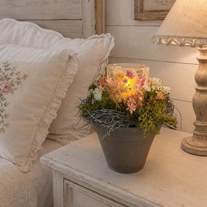 Peut inclure: Un pot en céramique grise contenant un bougeoir en verre avec une flamme chaude et lumineuse. La bougie est entourée d'un arrangement décoratif de mousse verte, de brindilles séchées et de petites fleurs artificielles roses, blanches et orange.
