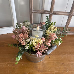 Könnte beinhalten: Ein kleiner, verzinkter Metalleimer gefüllt mit künstlichen Blumen in Rosa, Gelb und Weiß, um einen Glas-Kerzenhalter mit einer brennenden Kerze herum. Der Eimer hat einen Metallgriff und steht auf einer Holzoberfläche.