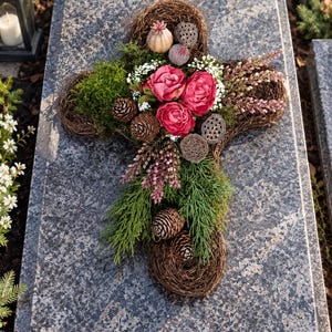 May include: A cross-shaped floral arrangement crafted from woven brown twigs, decorated with three deep pink roses, green moss, pine cones, dried lotus seed pods, small white flowers, and sprigs of pink heather, resting on a grey granite surface.