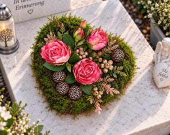 Puede incluir: Un arreglo floral en forma de corazón con rosas de color rosa intenso, pequeñas bayas verdes y piñas marrones sobre una base de musgo verde. El conjunto se encuentra sobre una piedra gris claro con el texto In liebevoller Erinnerung.