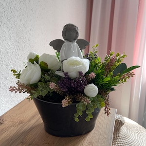Könnte beinhalten: Blumenarrangement in einem schwarzen Topf mit weißen Rosen, grünem Laub und lila Akzenten. Eine kleine graue Engelsfigur sitzt inmitten der Blumen. Das Arrangement soll eine ruhige und elegante Atmosphäre schaffen.