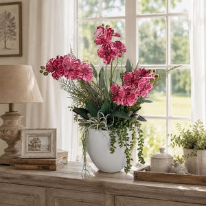 Op de afbeelding: Een witte, ronde keramische pot met een levendig arrangement van magenta en wit gespikkelde orchideeën, groene varenbladeren, hangende vetplantstengels en luchtplanten, wat zorgt voor een weelderige, botanische uitstraling.