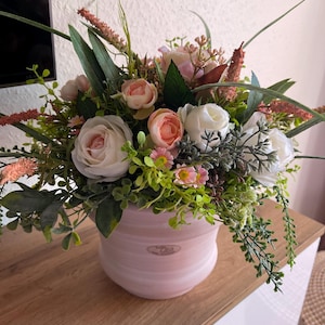 Könnte beinhalten: Blumenarrangement in einem rosa Keramiktopf. Die Anordnung umfasst weiße, rosa und pfirsichfarbene Rosen sowie grünes Laub und kleine rosa Blüten. Der Topf hat einen Marmoreffekt und das Wort "Casa" ist sichtbar.