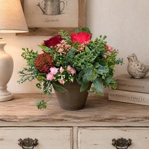 Op de afbeelding: Een bloemstuk in een donkergrijze pot met dieprode rozen, een donkerrode dahlia, kleine roze bloesems en diverse groene bladeren. Het arrangement staat op een lichtgekleurd houten oppervlak met twee lades en decoratieve metalen handgrepen.