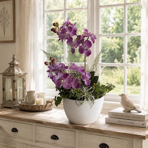 Può includere: Una composizione di orchidee artificiali in vaso con petali viola maculati su steli alti, arricchita da fogliame verde e piante aeree, contenuta in un vaso bianco opaco a forma di ciotola, posizionata su una superficie in legno.