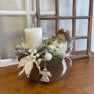 Op de afbeelding: Een decoratief middelpunt in een bruine kom, met een grote witte kaars, bevroren groen, witte ornamenten en een klein engelenbeeldje. Een houten engelenornament hangt aan de kom. De opstelling staat voor een raam.