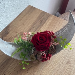 Può includere: Un centrotavola decorativo con una vibrante rosa rossa, circondata da verde e bacche, in un contenitore grigio a forma di foglia. La composizione è posta su una superficie di legno.