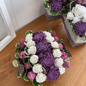 Puede incluir: Arreglo floral con rosas blancas y flores moradas texturizadas. El arreglo incluye pequeñas piñas y está colocado en una cesta tejida. El follaje verde y blanco acentúa las flores.