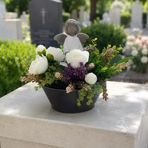 Può includere: Una ciotola nera riempita con una composizione di fiori bianchi simili a peonie, piccoli grappoli viola e fogliame verde. Una piccola statuetta di angelo grigio con vesti bianche è posizionata al centro della composizione floreale.