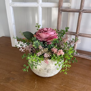 Könnte beinhalten: Ein dekoratives Blumengesteck in einem runden, strukturierten, steinähnlichen Topf. Das Arrangement zeigt eine große, rosa Rose, umgeben von grünem und rosa Laub. Der Topf steht auf einer Holzoberfläche, mit einem Fenster und einem Holzrahmen im Hintergrund.