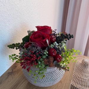 Könnte beinhalten: Dekorative Blumengesteck mit zwei großen roten Rosen, roten Beeren und grünem Laub. Die Anordnung befindet sich in einem strukturierten, grauen Keramiktopf. Ideal für die Inneneinrichtung oder als Geschenk.