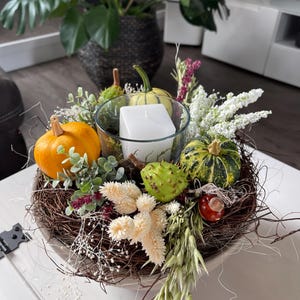 Può includere: Un centrotavola decorativo con un portacandele in vetro trasparente e una candela bianca quadrata, circondato da piccole zucche, un riccio di castagna verde spinoso, fiori bianchi secchi, fogliame verde e ramoscelli marroni in cerchio.