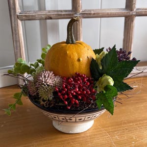 Op de afbeelding: Een decoratieve kom met een witte voet en zwarte geometrische patronen op de rand, gevuld met een centrale oranje pompoen, groen blad, kleine roze getextureerde clusters, felrode besachtige takjes en donkerpaarse accenten.
