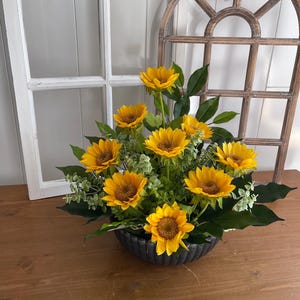 Puede incluir: Un arreglo floral con girasoles amarillos brillantes con centros marrones, hojas verdes y pequeñas flores blancas. Los girasoles están dispuestos en un cuenco gris oscuro texturizado. El arreglo está sobre una superficie de madera.