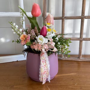 Puede incluir: Arreglo floral en una maceta de color lila. El arreglo incluye flores rosas, blancas y amarillas, con follaje verde. Una cinta con un estampado floral y ribete de encaje adorna la maceta. El fondo presenta un marco de ventana de madera.