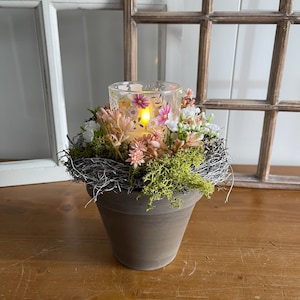 Può includere: Un portacandele decorativo con un lumino in vetro con disegni floreali, circondato da un nido di ramoscelli secchi, muschio e fiori. Il portacandele è posto in un vaso di ceramica marrone, creando un'estetica naturale e rustica.