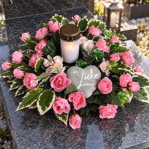 Könnte beinhalten: Ein Blumenkranz mit rosa Rosen und panaschierten grünen Blättern, der eine weiße Stumpenkerze und einen grauen herzförmigen Stein mit dem weißen Schriftzug "in Liebe" auf der Vorderseite umschließt, platziert auf einer dunklen Granitplatte.